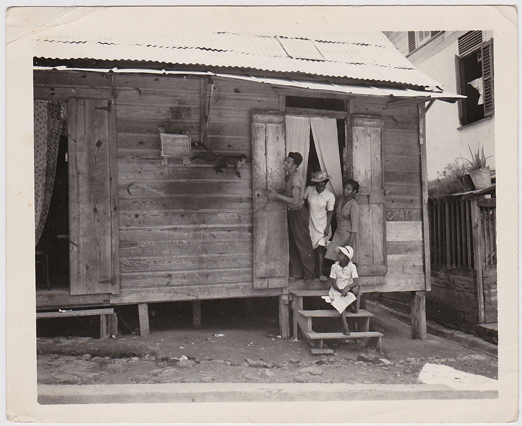 A strange image of three black women and a white man with a cigarette at the door of a simple wood house, watching a monkey climb towards them. 