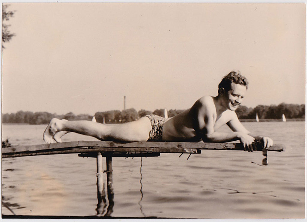 Reclining on a diving board, wearing a sort of leopard skin bathing suit and holding sunglasses in his hand, a handsome young guy smiles for the camera.