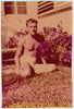 Very early color snapshot of a handsome blond man wearing shorts, sitting on the lawn next to a bed of poinsettias. It has the golden yellow hue typical of early color photos and the je ne sais quoi that tilts "gay."