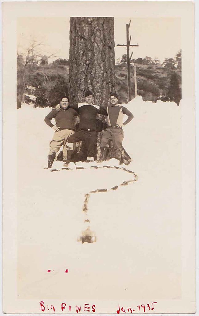 Three guys strike a pose above a question mark made up of what looks like beer bottles in the snow. Vintage snapshot