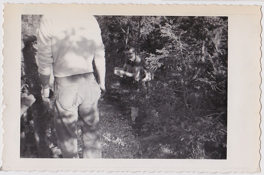 Anonymous vintage snapshot of a smiling man crouching (defecating) in the woods with an audience of at least two.
