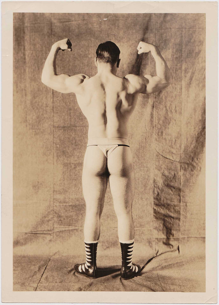 Vintage sepia photo strongman wearing a posing strap and fancy wrestling boots, flexing his biceps, gay