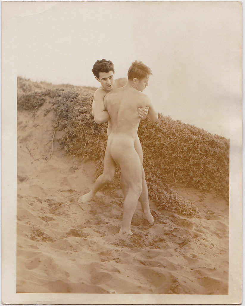 Two handsome young nude models wrestle in the sand.